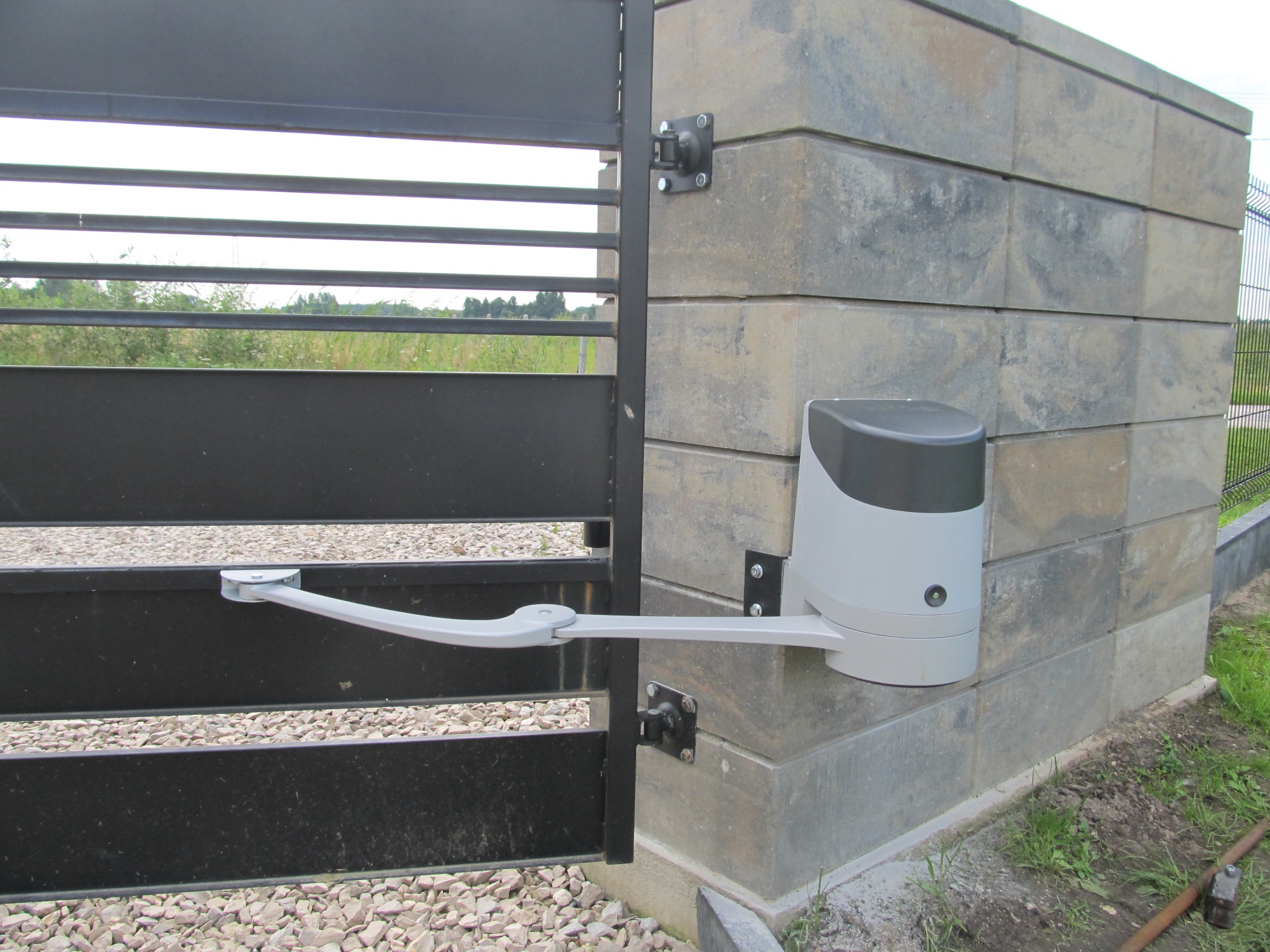 Szara, pozioma brama z automatycznym napędem zamontowanym na słupku z szarej kostki, widok z bliska na mechanizm otwierający.