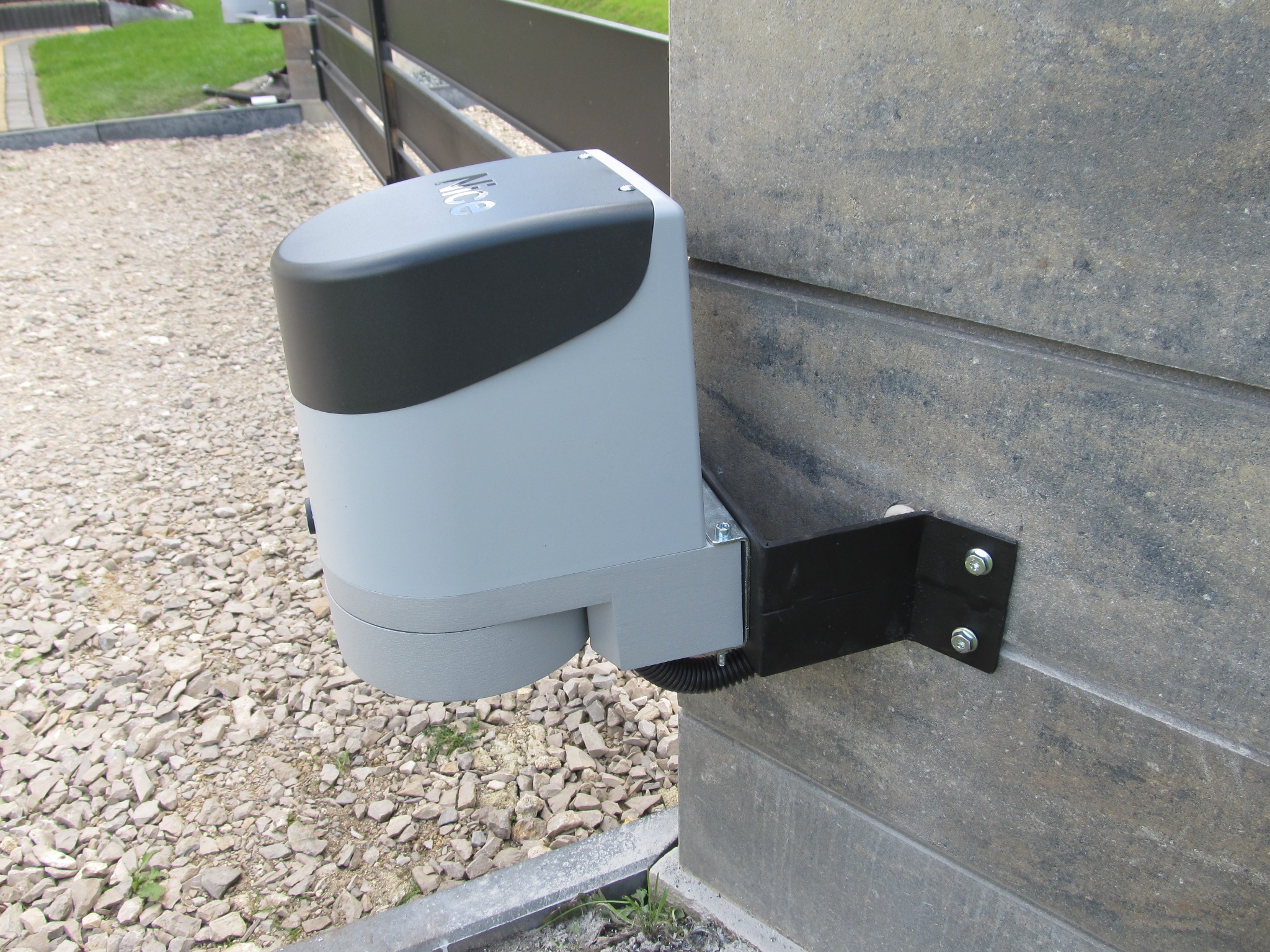 Szary napęd do bramy zamontowany na kamiennym słupku, widoczne śruby mocujące i fragment ogrodzenia.