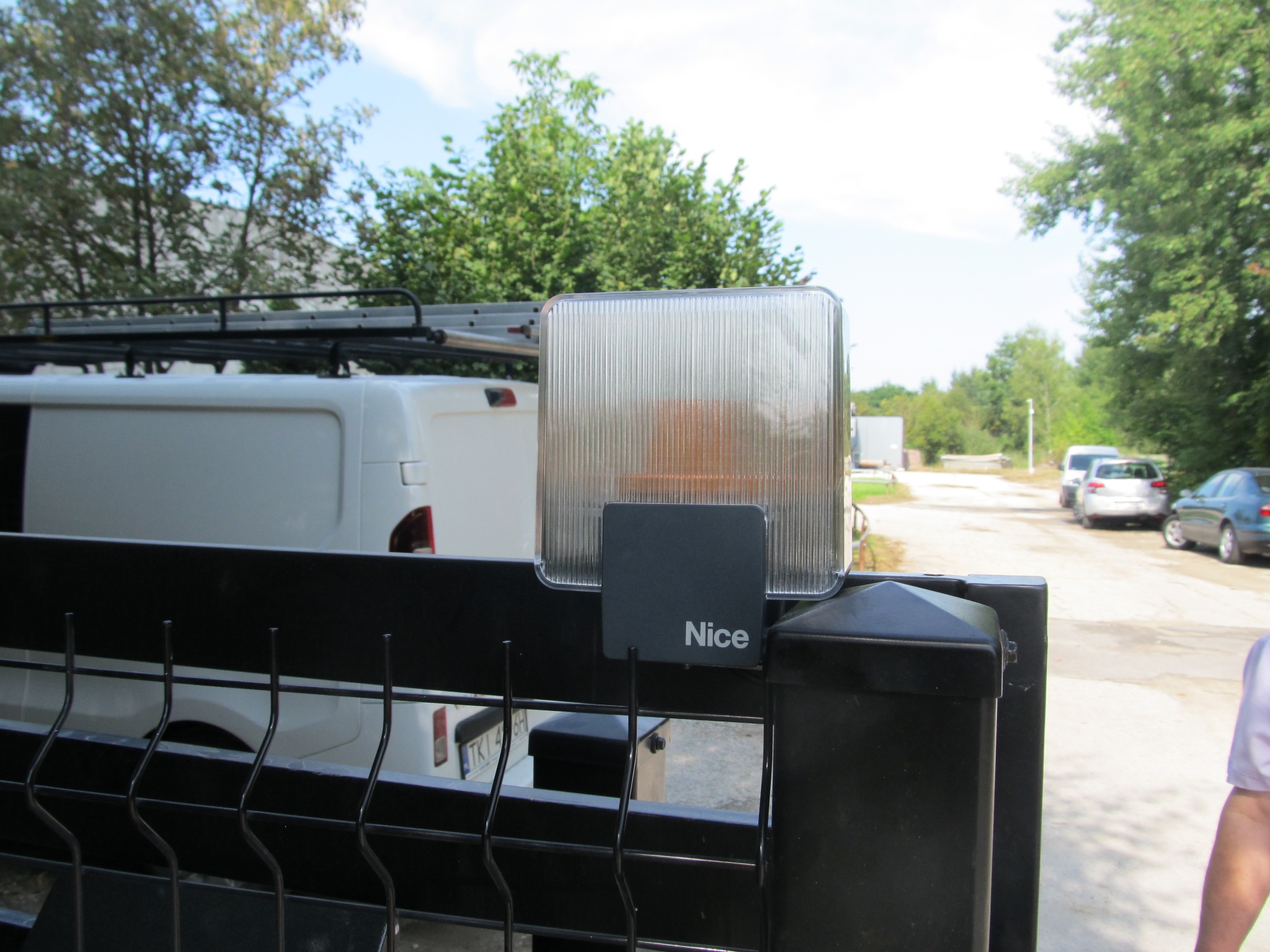 Lampa ostrzegawcza i tabliczka z logo Nice na czarnym słupku bramy wjazdowej, w tle biały bus z drabinką na dachu i inne zaparkowane pojazdy.