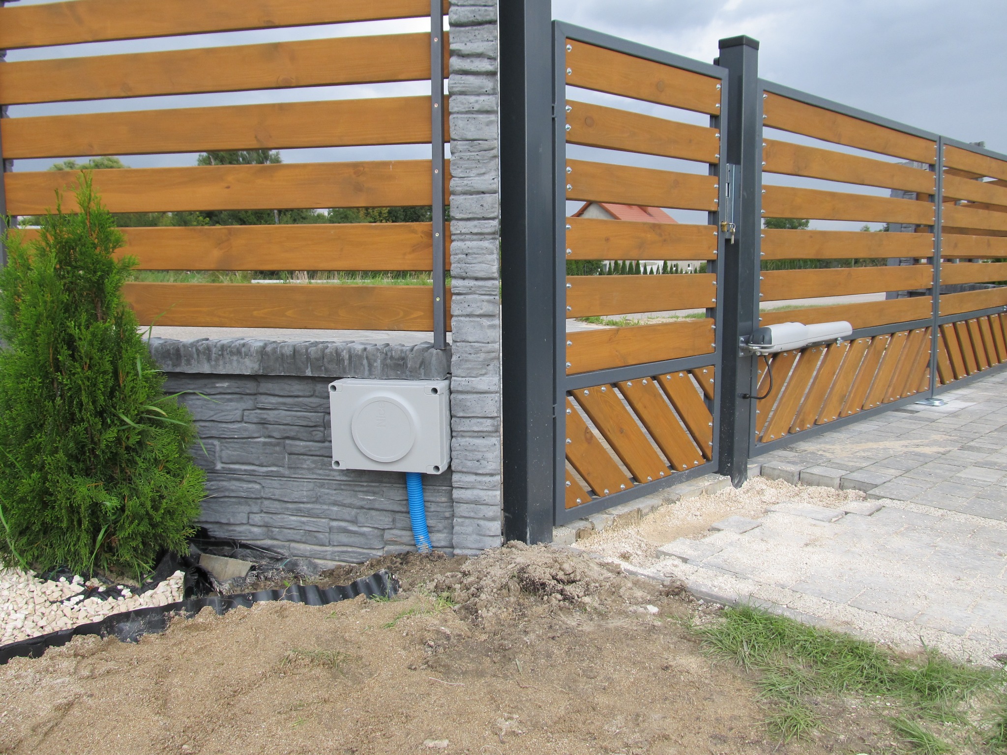 Nowoczesna brama wjazdowa z drewnianymi panelami i automatyką, widoczna skrzynka sterownicza z niebieskim peszlem, świeżo obsypana ziemią.