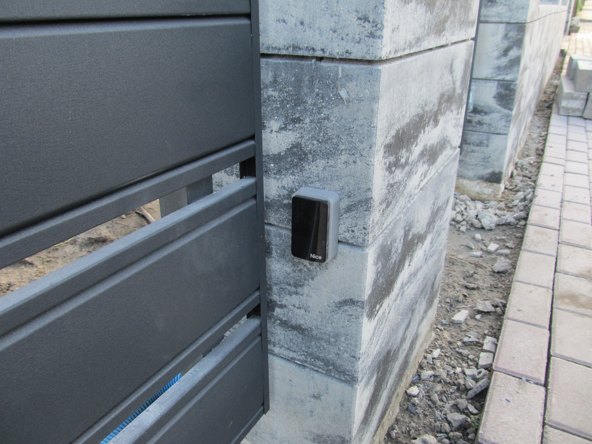 Szara brama z automatyką Nice zamontowaną na betonowym słupku z szarej kostki, widoczne elementy instalacji w szczelinie między bramą a słupkiem.
