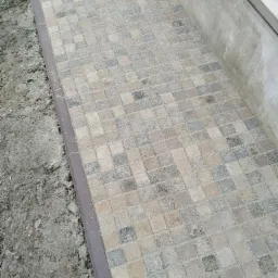 Ułożona ścieżka z kostki brukowej o kwadratowym kształcie w odcieniach szarości i beżu, obramowana brązowym obrzeżem, przylegająca do betonowego murku i ziemi.