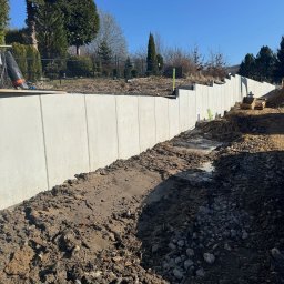 LITBUD Mateusz Litewka - Wysoka, betonowa ściana oporowa w trakcie budowy, widoczne ślady ziemi i kamieni, na górze siatka zabezpieczająca, w tle drzewa i błękitne niebo.