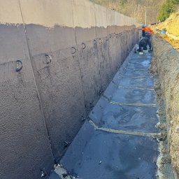 LITBUD Mateusz Litewka - Hydroizolacja betonowego koryta odpływowego, widoczni dwaj pracownicy w odzieży roboczej, teren pofałdowany w tle.