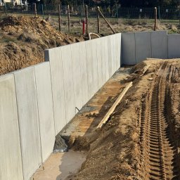 LITBUD Mateusz Litewka - Nowo wzniesiona, długa, szara ściana oporowa z betonu, widoczne ślady opon ciężkiego sprzętu na ziemi i drewniane elementy szalunku, w tle dom z panelami słonecznymi i drzewa.