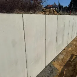 Wzniesiony mur oporowy z prefabrykatów betonowych, widoczny młotek i fragment deski, w tle ziemia i zabudowa jednorodzinna.