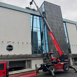 LITBUD Mateusz Litewka - Czerwony podnośnik teleskopowy Manitou MRT podnosi pakiet materiałów na dach budynku z szarą elewacją, widoczny również czerwony samochód dostawczy i drabina.