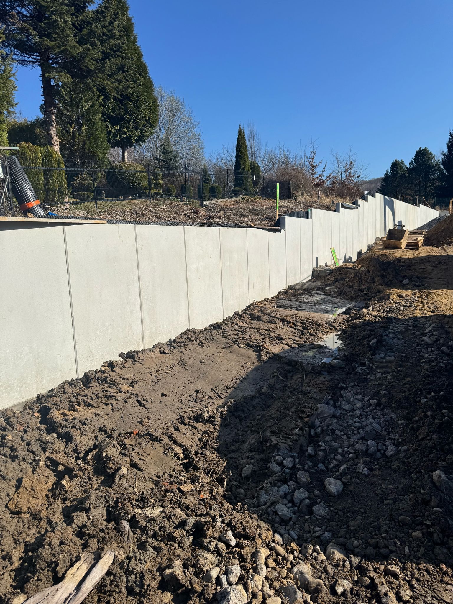 Wysoka, betonowa ściana oporowa w trakcie budowy, widoczne ślady ziemi i kamieni, na górze siatka zabezpieczająca, w tle drzewa i błękitne niebo.