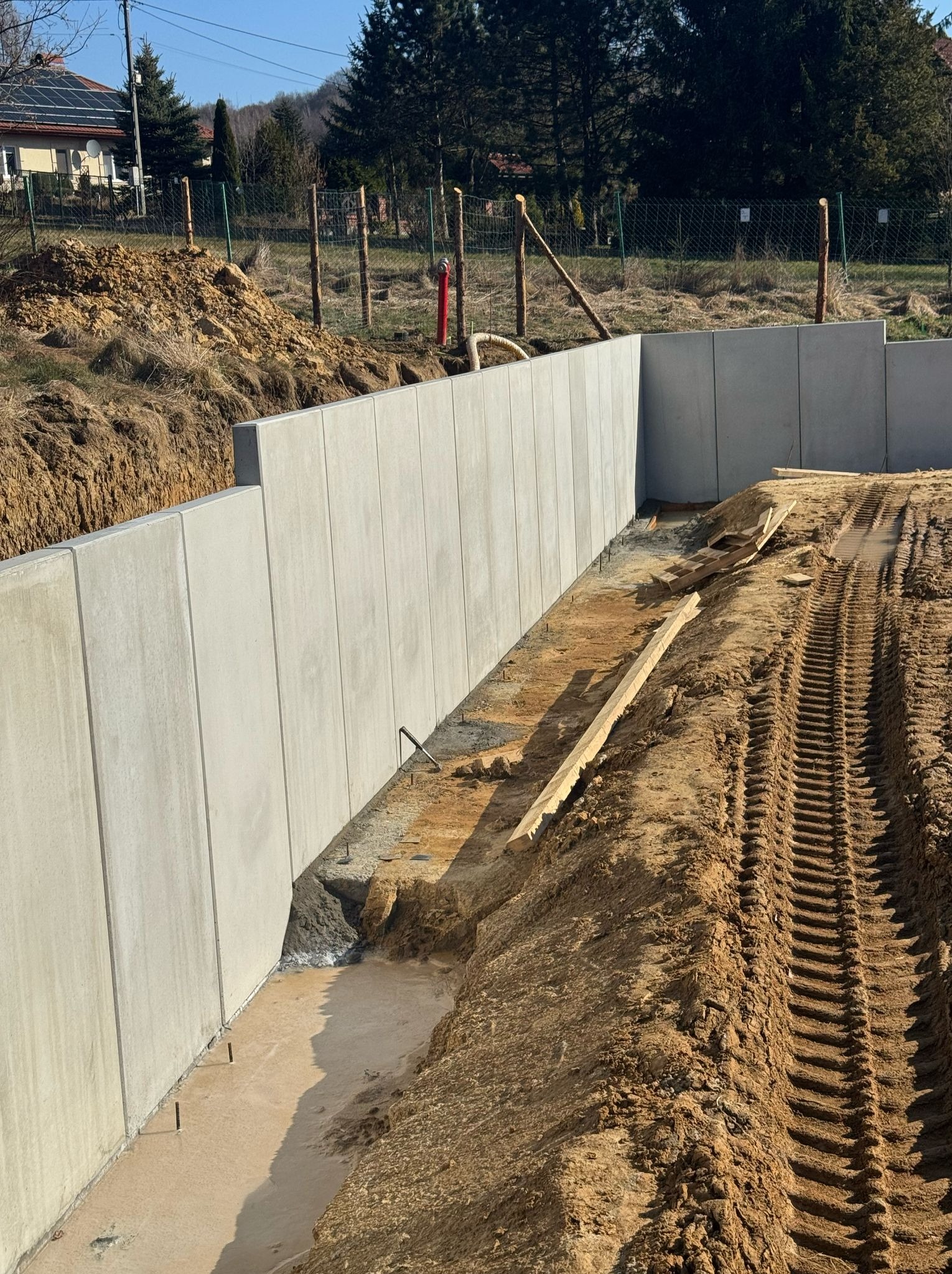 Nowo wzniesiona, długa, szara ściana oporowa z betonu, widoczne ślady opon ciężkiego sprzętu na ziemi i drewniane elementy szalunku, w tle dom z panelami słonecznymi i drzewa.