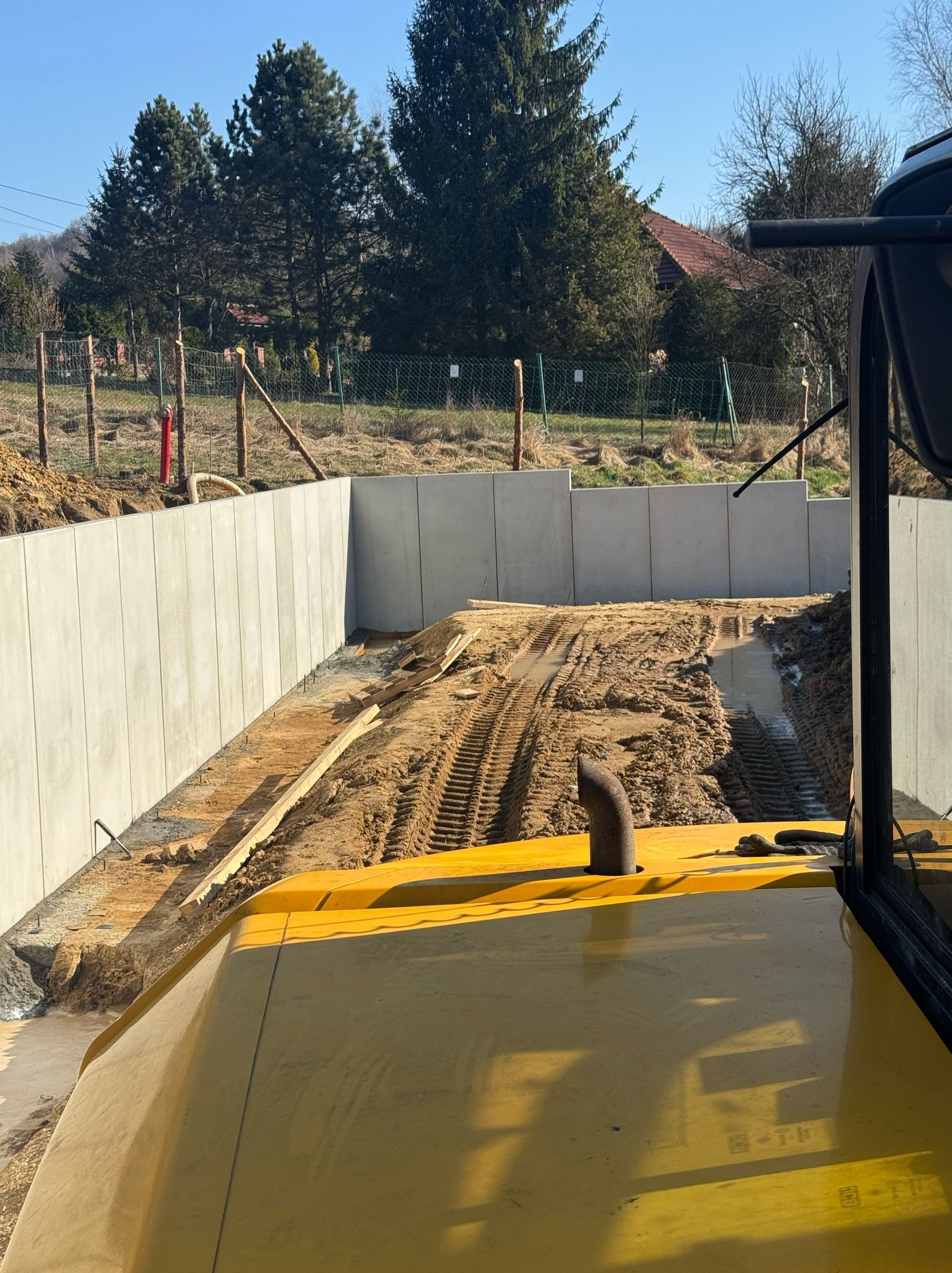 Widok z kabiny żółtej maszyny budowlanej na wykop z betonowymi ścianami oporowymi i śladami opon w błotnistej ziemi, w tle ogrodzenie i drzewa.