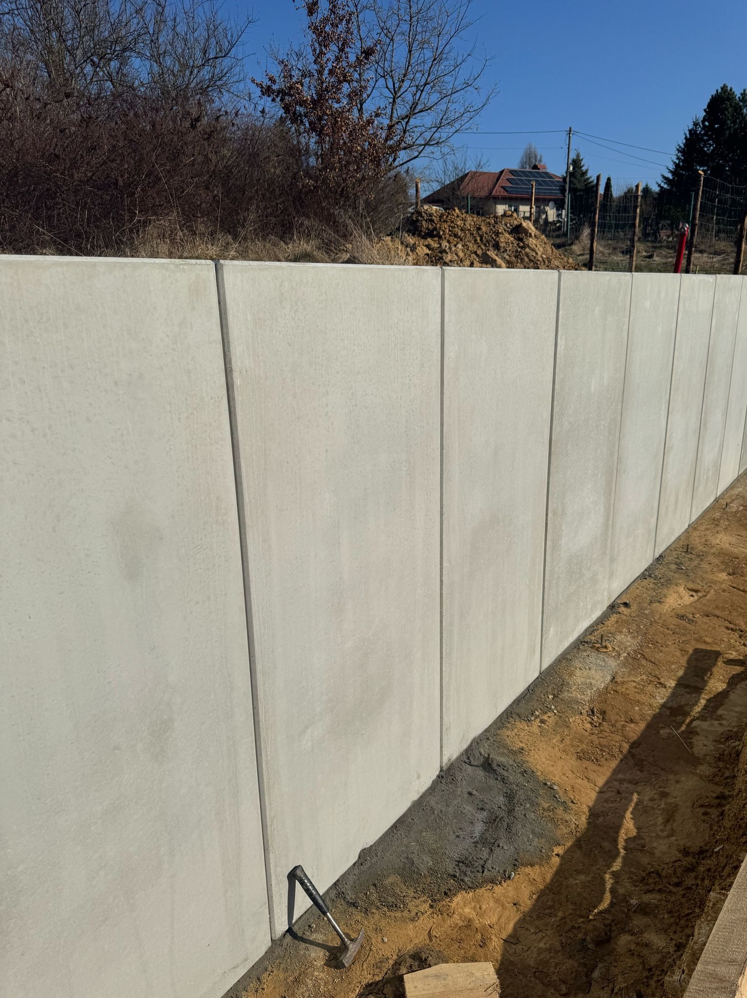 Wzniesiony mur oporowy z prefabrykatów betonowych, widoczny młotek i fragment deski, w tle ziemia i zabudowa jednorodzinna.
