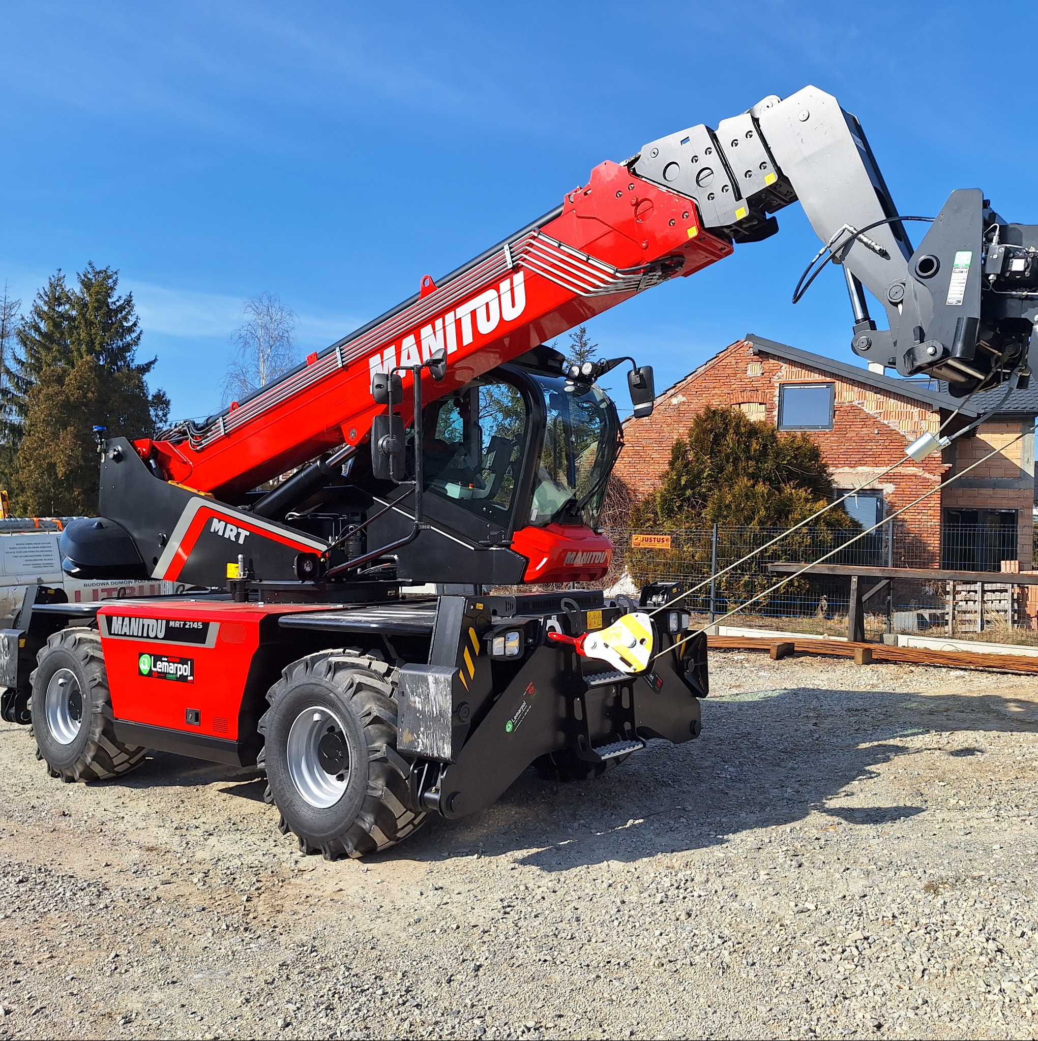 Czerwony teleskopowy podnośnik Manitou MRT 2145 na placu budowy, w tle budynek z czerwonej cegły w trakcie wznoszenia.