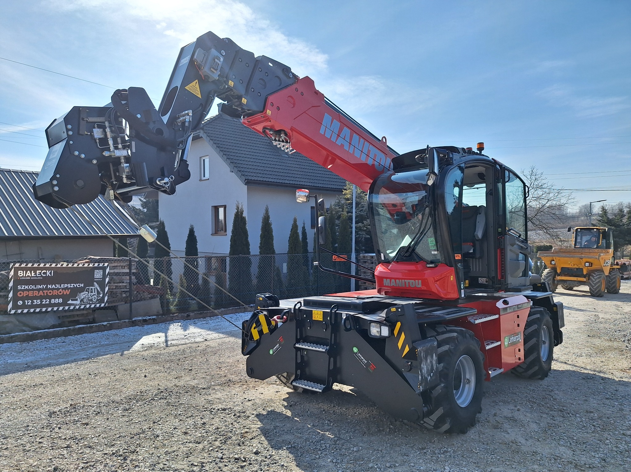 Czerwony telehandler Manitou MRT 2145 z osprzętem roboczym na tle domu jednorodzinnego i tablicy reklamowej szkoły operatorów maszyn budowlanych.