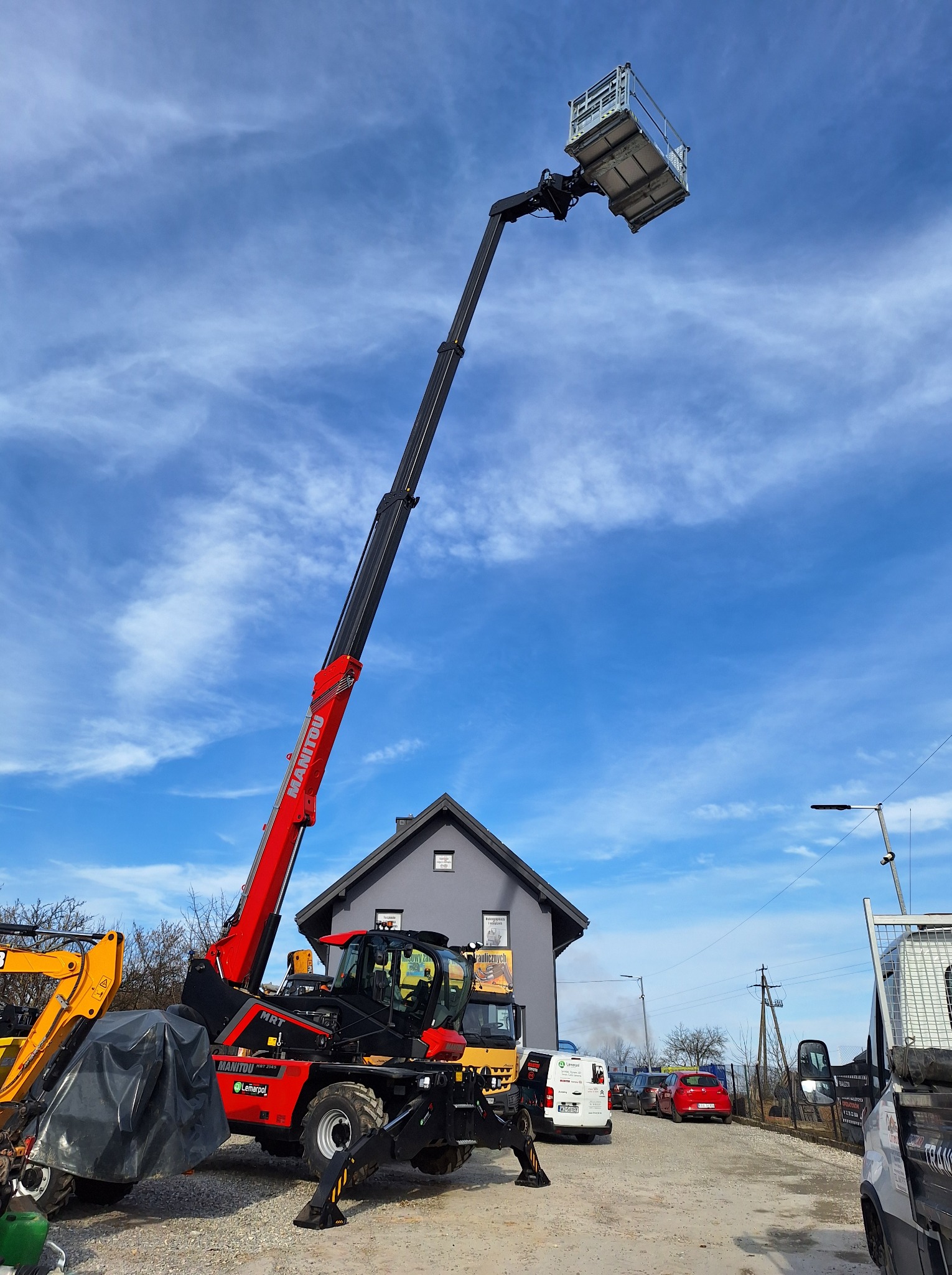 Czerwono-czarny podnośnik teleskopowy Manitou z wysuniętym ramieniem i platformą roboczą na tle błękitnego nieba z chmurami, zaparkowany na żwirowym placu budowy z widocznymi innymi maszynami...