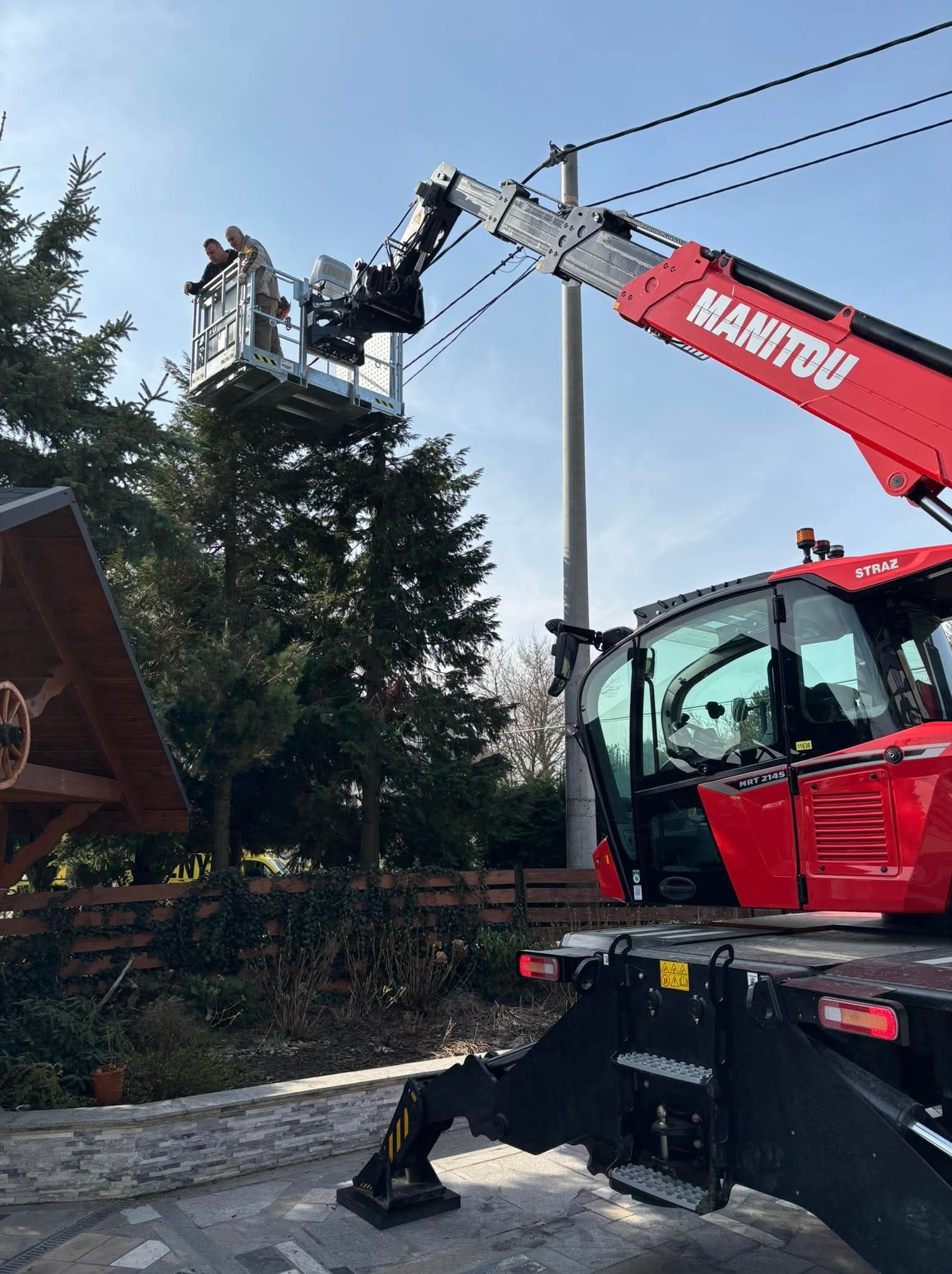 Dwóch pracowników w koszu podnośnika Manitou, pracujących przy linii energetycznej, widoczny fragment dachu altany i drzewa.