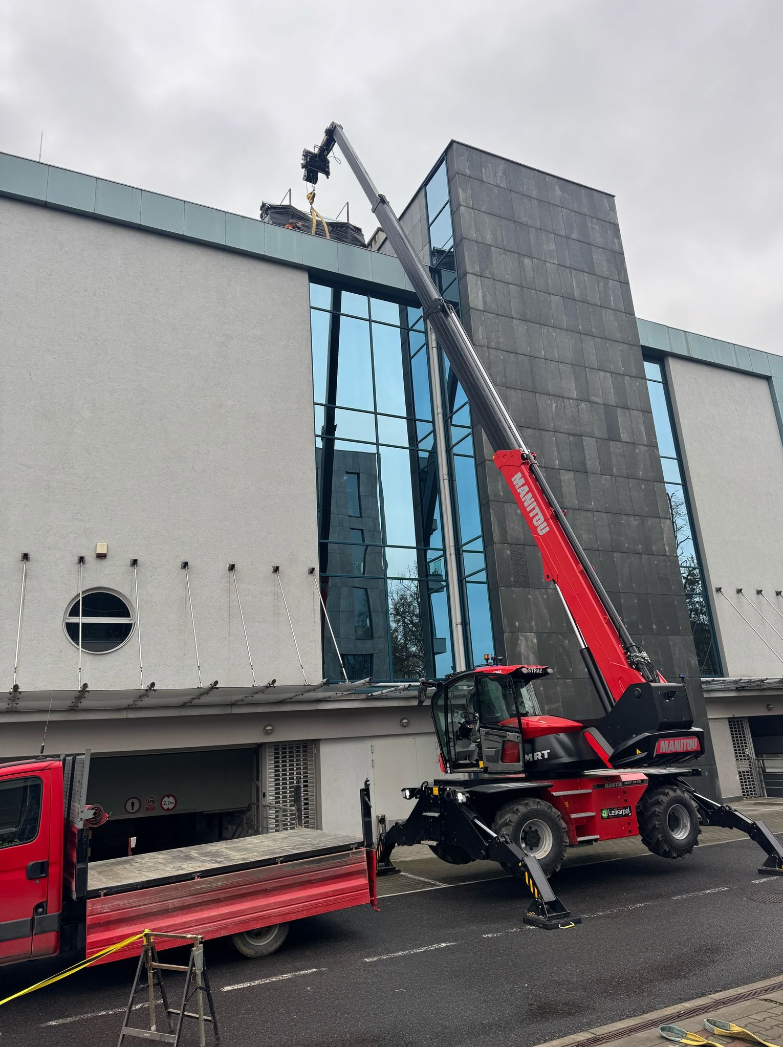 Czerwony podnośnik teleskopowy Manitou MRT podnosi pakiet materiałów na dach budynku z szarą elewacją, widoczny również czerwony samochód dostawczy i drabina.