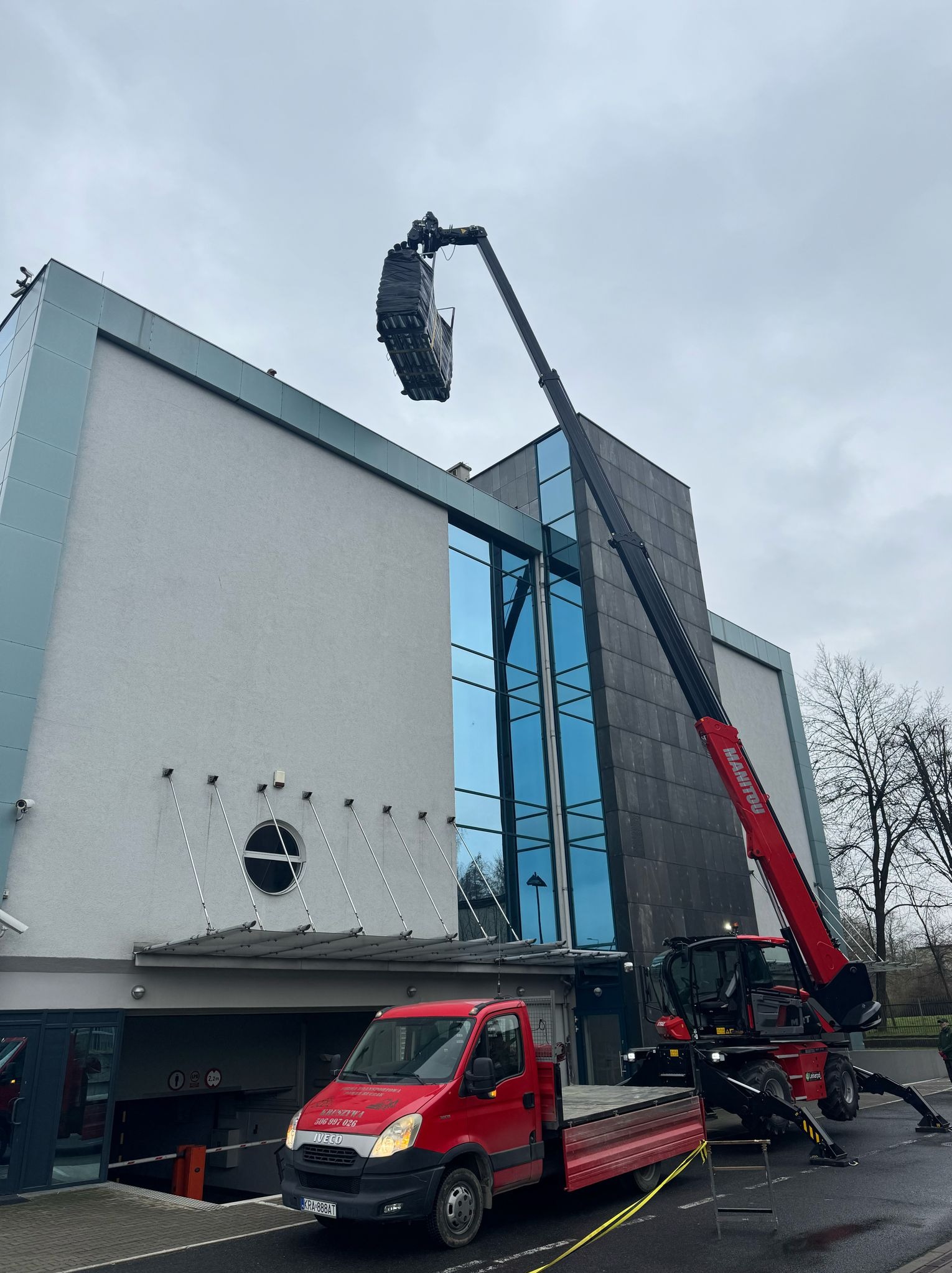 Czerwony dźwig teleskopowy Manitou podnosi paletę z materiałami budowlanymi na wysokość budynku z szarą elewacją i niebieskimi oknami, obok stoi czerwony samochód dostawczy Iveco.