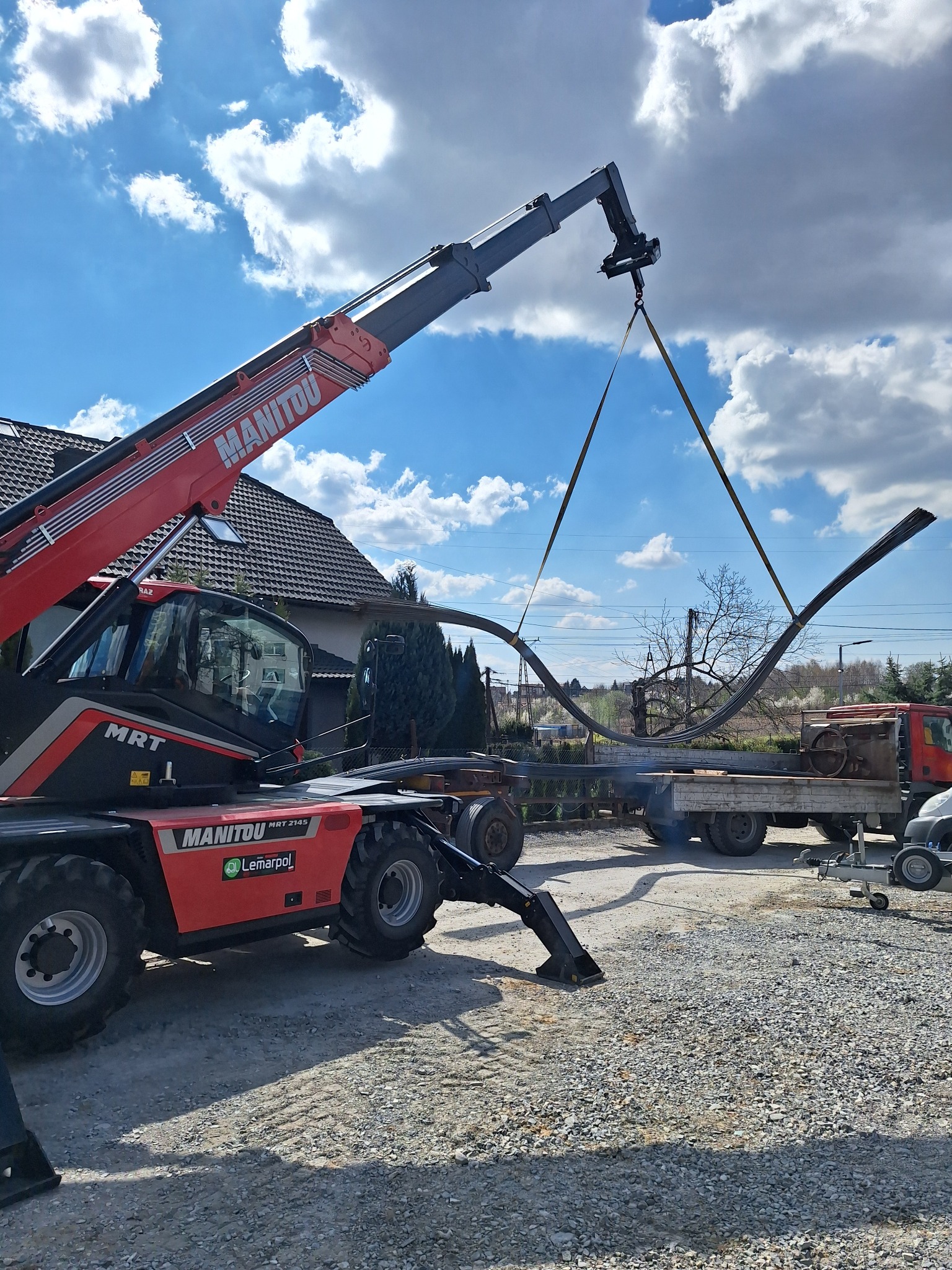 Czerwony podnośnik teleskopowy Manitou transportuje stalowe pręty zbrojeniowe na tle błękitnego nieba z chmurami w Giebułtowie.