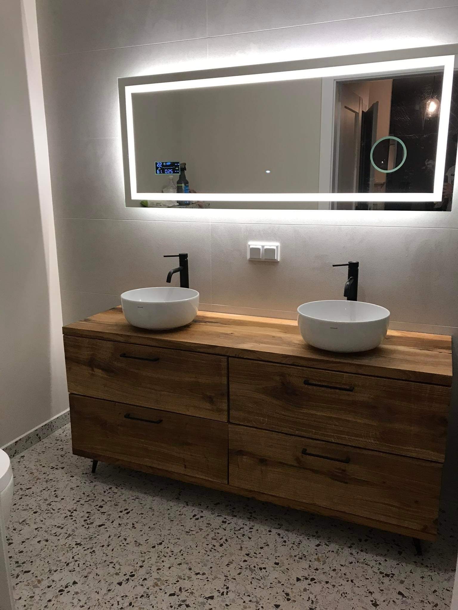 Nowoczesna łazienka z drewnianą szafką pod umywalki nablatowe, czarne baterie, podświetlane lustro LED i podłoga lastryko.