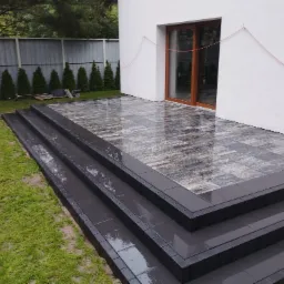 Nowoczesny taras z kostki brukowej w odcieniach szarości, prowadzący do drzwi balkonowych, z widocznymi trzema stopniami i schludnym trawnikiem w tle.