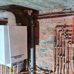 Montaż instalacji grzewczej z piecem gazowym: widoczne miedziane rury, zawory i szara rura odprowadzająca spaliny, zamontowane na ceglanej ścianie w pomieszczeniu o surowym, betonowym suficie.