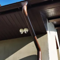 Dwa białe kamery monitoringu kopułkowe zamontowane pod brązowym okapem domu, widok z dołu na tle fragmentu nieba z chmurami.