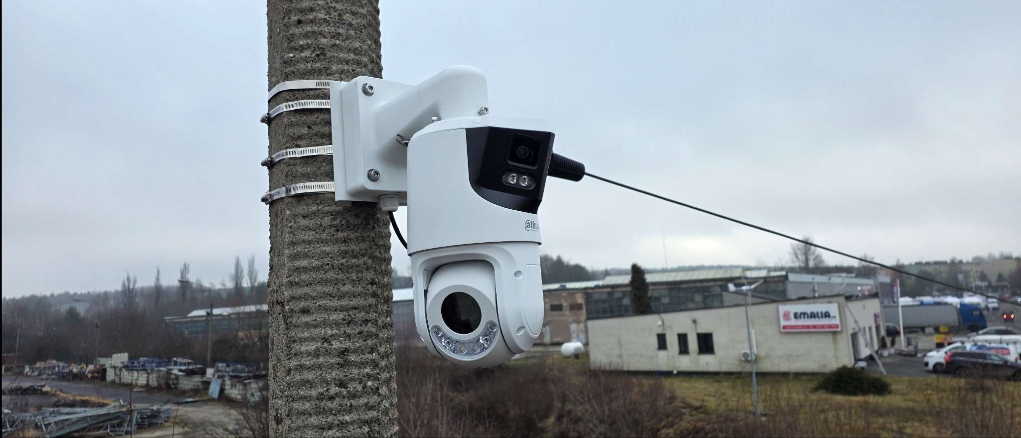 Kamera PTZ Dahua zamontowana na betonowym słupie za pomocą metalowych obejm. W tle budynki przemysłowe i pochmurne niebo. Widok z bliska na zainstalowany system monitoringu.