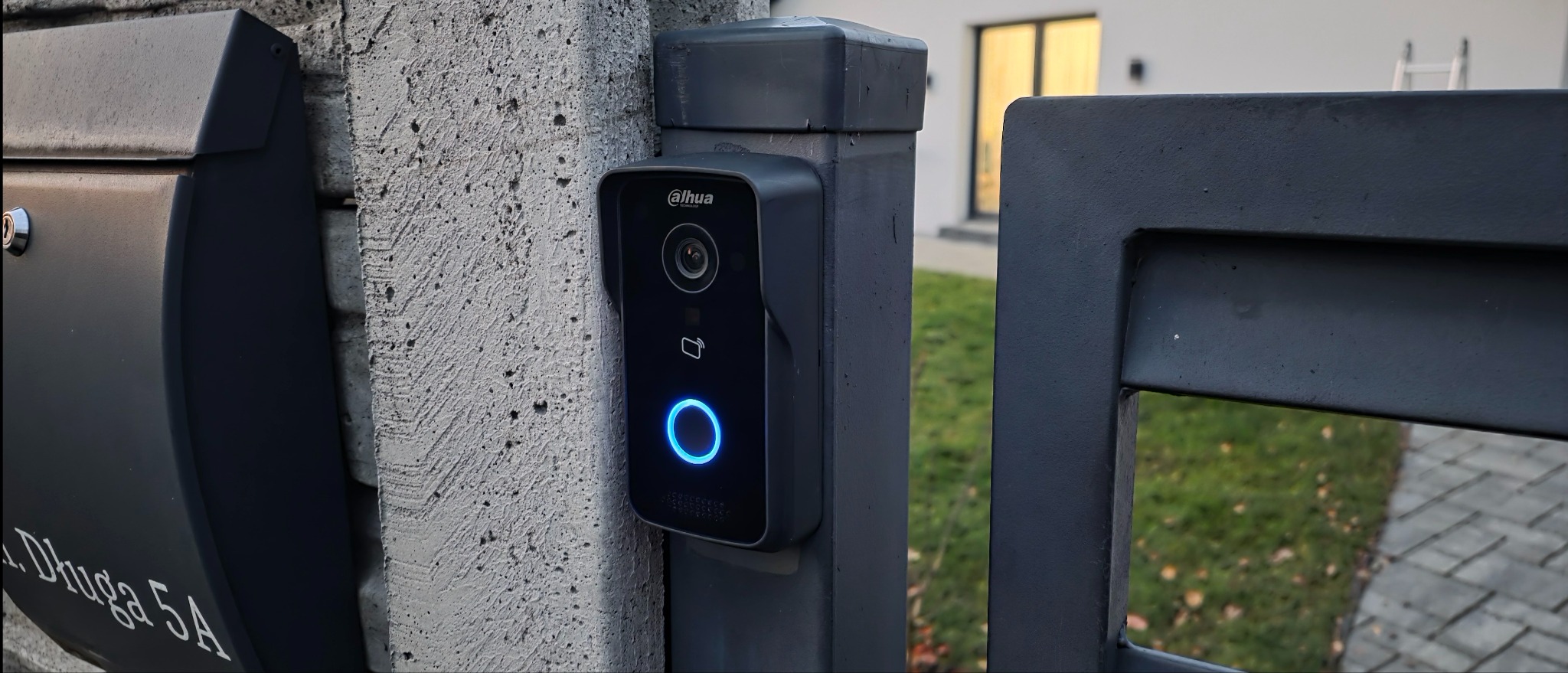 Nowoczesny wideodomofon Dahua zamontowany na betonowym słupku obok skrzynki pocztowej i metalowej bramy. Widoczny podświetlany przycisk i kamera. W tle dom z oknami.
