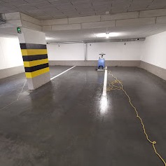 Błyszcząca, mokra posadzka w garażu podziemnym po czyszczeniu, widoczna maszyna czyszcząca z długim, zwiniętym kablem, słup oznaczony czarno-żółtymi pasami.