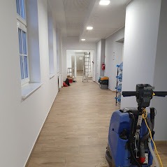 Automat szorujący podłogę w długim korytarzu z jasnymi ścianami i drewnopodobną podłogą. W tle widoczne drabiny i butelki z wodą.