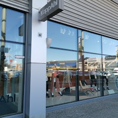 Widok z zewnątrz na witrynę sklepu KappaAhl, z wyraźnie odbijającym się niebem i budynkami w szybach. Widoczny chodnik i fragment elewacji budynku.