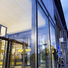 Wysokie okna budynku komercyjnego po umyciu, widoczne odbicia w szybach, drabina aluminiowa z wiadrem i ściągaczką w pobliżu.