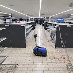 Automat szorujący podłogę w pustym sklepie z regałami, widoczne kable i węże, jasne oświetlenie sufitowe.