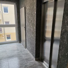 Wnętrze klatki schodowej z widocznym oknem, drzwiami i windą, ściany pokryte ciemnym, wzorzystym tynkiem.