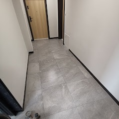 Wypolerowana, szara podłoga z płytek w korytarzu mieszkania, widoczne ślady po maszynie polerskiej w lewym dolnym rogu, jasne ściany i drzwi wejściowe.