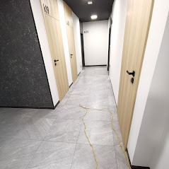 Korytarz w nowoczesnym apartamentowcu z szarymi płytkami podłogowymi, jasnymi drzwiami z czarnymi klamkami i numerami pokoi, widoczne ślady po remoncie w postaci zabrudzeń na podłodze.