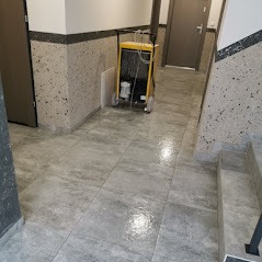 Profesjonalny sprzęt do czyszczenia posadzek na kółkach w korytarzu wyłożonym szarymi płytkami imitującymi kamień, widoczne ślady wilgoci po czyszczeniu.