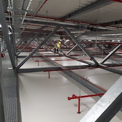 Pracownik w żółtym kombinezonie i kasku, zabezpieczony liną, porusza się po stalowej konstrukcji pod sufitem hali przemysłowej z widocznymi instalacjami przeciwpożarowymi.