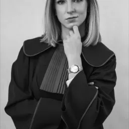 Portret kobiety-adwokata w todze, z ręką pod brodą i zegarkiem na nadgarstku, na neutralnym tle.