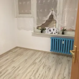 Pusty pok&oacute;j po remoncie z jasnymi panelami podłogowymi ułożonymi wzdłuż pokoju, błękitny grzejnik pod oknem z dekoracją 'LOVE' i kwiatami.