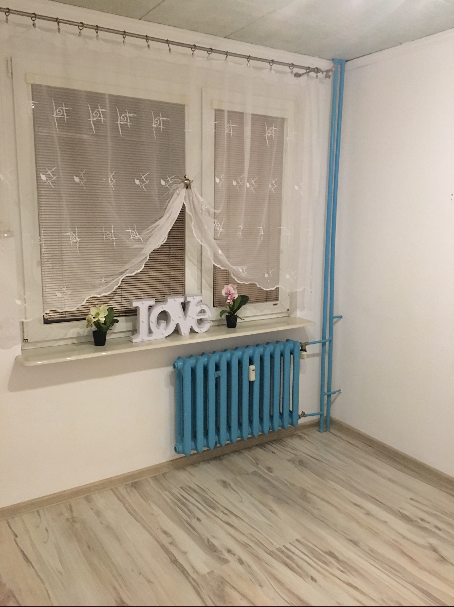 Pusty pokój z jasnymi panelami podłogowymi, błękitnym grzejnikiem i rurami, okno z firankami i roletami, na parapecie dekoracja LOVE i kwiatki.