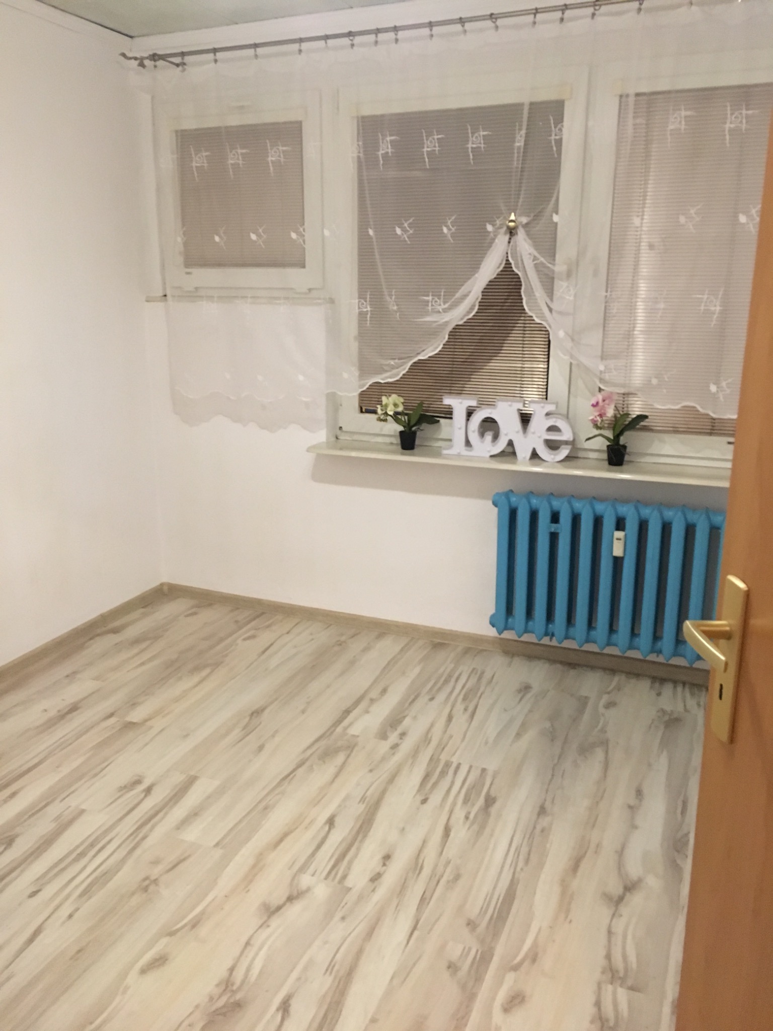Pusty pokój po remoncie z jasnymi panelami podłogowymi ułożonymi wzdłuż pokoju, błękitny grzejnik pod oknem z dekoracją 'LOVE' i kwiatami.
