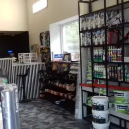 Wnętrze sklepu budowlanego z regałami pełnymi farb, klejów i innych materiałów wykończeniowych, widoczny blat obsługi klienta z zegarem na ścianie w tle.