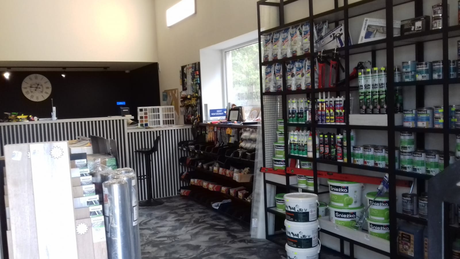 Wnętrze sklepu budowlanego z regałami pełnymi farb, klejów i innych materiałów wykończeniowych, widoczny blat obsługi klienta z zegarem na ścianie w tle.