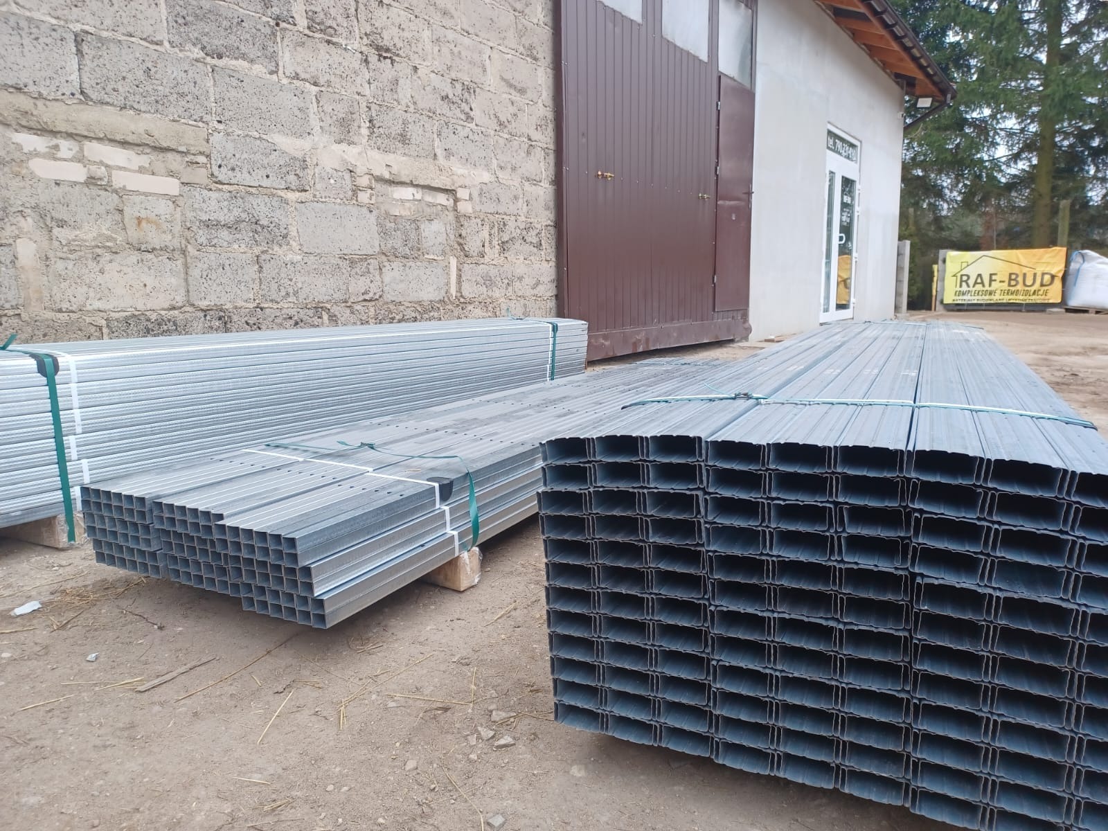 Stosy ocynkowanych profili metalowych C, spięte taśmą, składowane na placu budowy przed budynkiem z cegły i brązową bramą garażową.