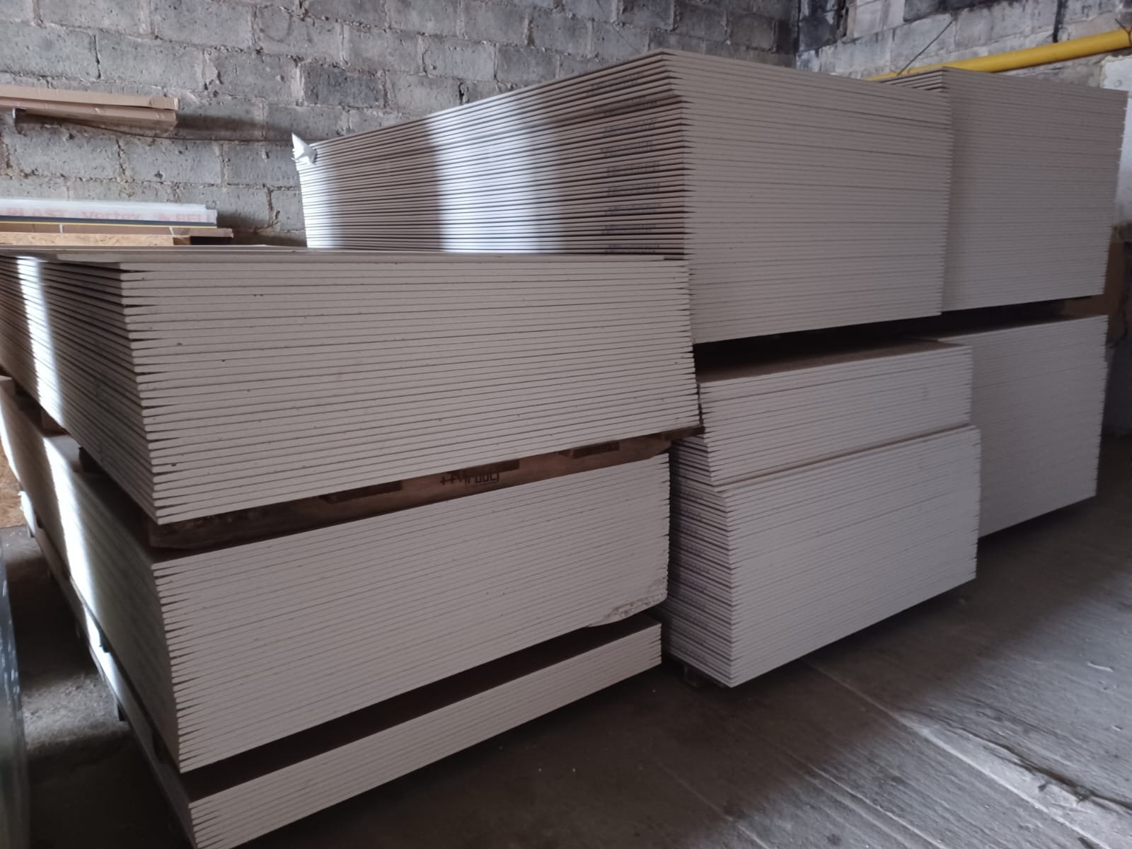 Stosy płyt gipsowo-kartonowych na paletach w magazynie o surowych, ceglanych ścianach. Widoczne uszkodzenia krawędzi płyt.