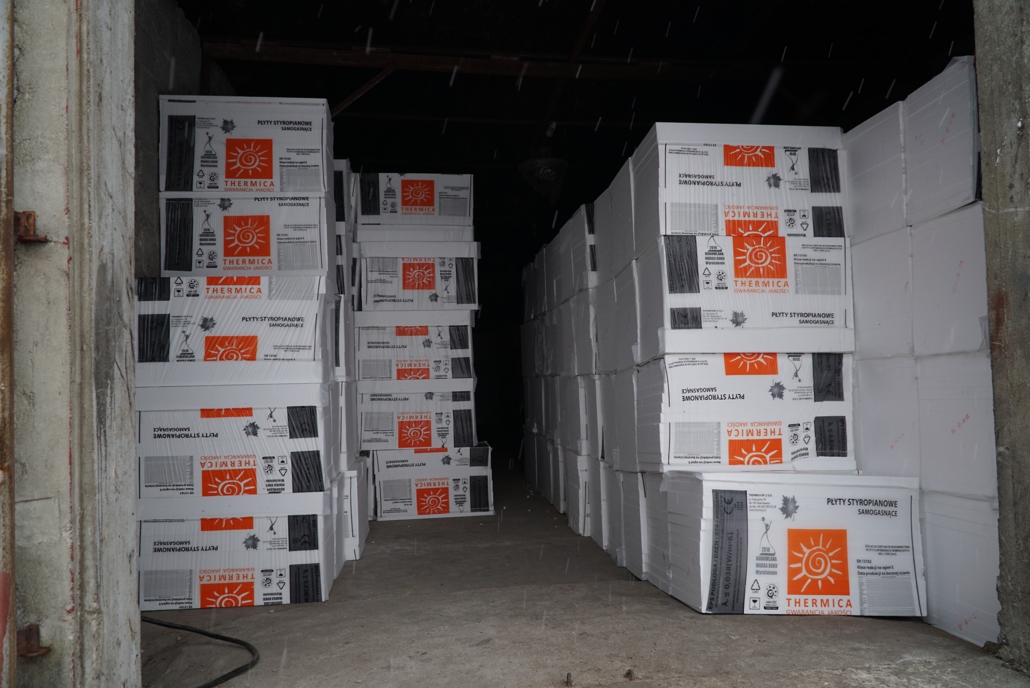 Magazyn z ułożonymi w stosy paczkami płyt styropianowych marki Thermica, widoczne etykiety informacyjne i logo producenta na opakowaniach.