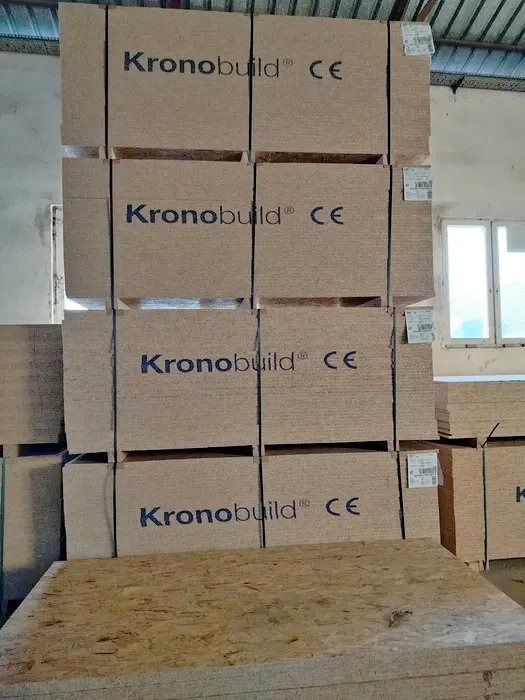 Stos płyt OSB Kronobuild z oznaczeniem CE, ułożonych w magazynie, widoczne etykiety i okno w tle.