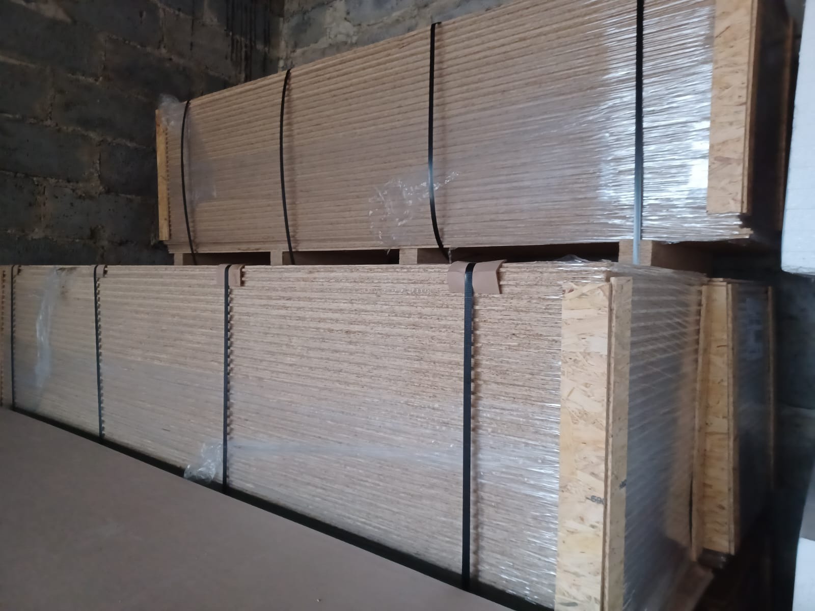 Dwa stosy płyt OSB, owinięte w folię i związane czarną taśmą, ułożone na paletach w magazynie z ceglanymi ścianami.