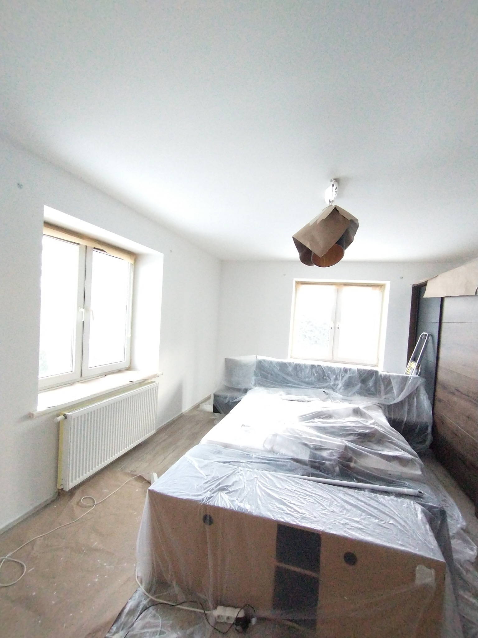 Pokój w trakcie remontu: świeżo pomalowane białe ściany, zabezpieczone meble folią malarską, zakryta lampa, widoczne ślady farby na podłodze.